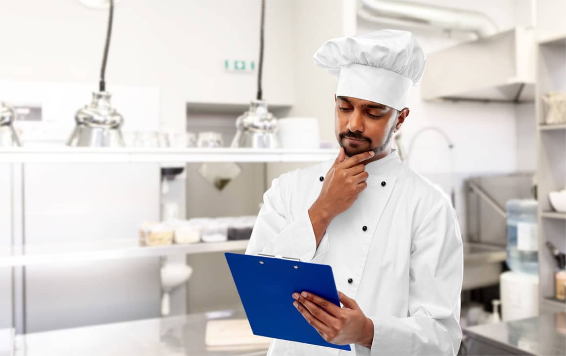A uniformed chef in a clean white kitchen, holding and looking at a clipboard while stroking his chin, looking thoughtful 